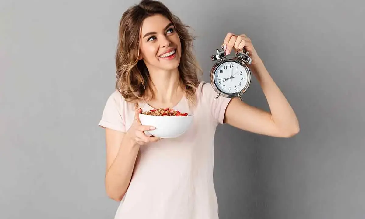 Mujer sonriente con un tazón de cereal y un despertador, lista para el ayuno intermitente para adelgazar 10 kilos.