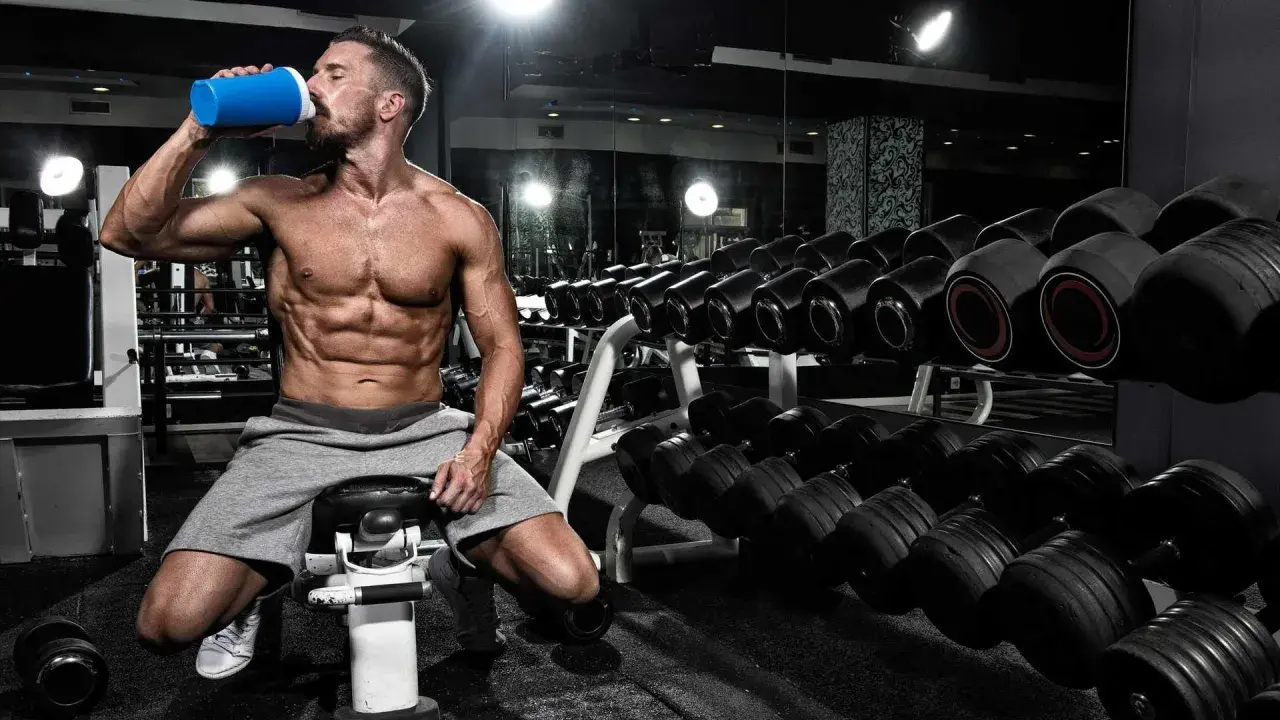 Hombre musculoso bebe de un batido azul en el gimnasio, rodeado de pesas. Es el momento perfecto para la ventana anabolica.