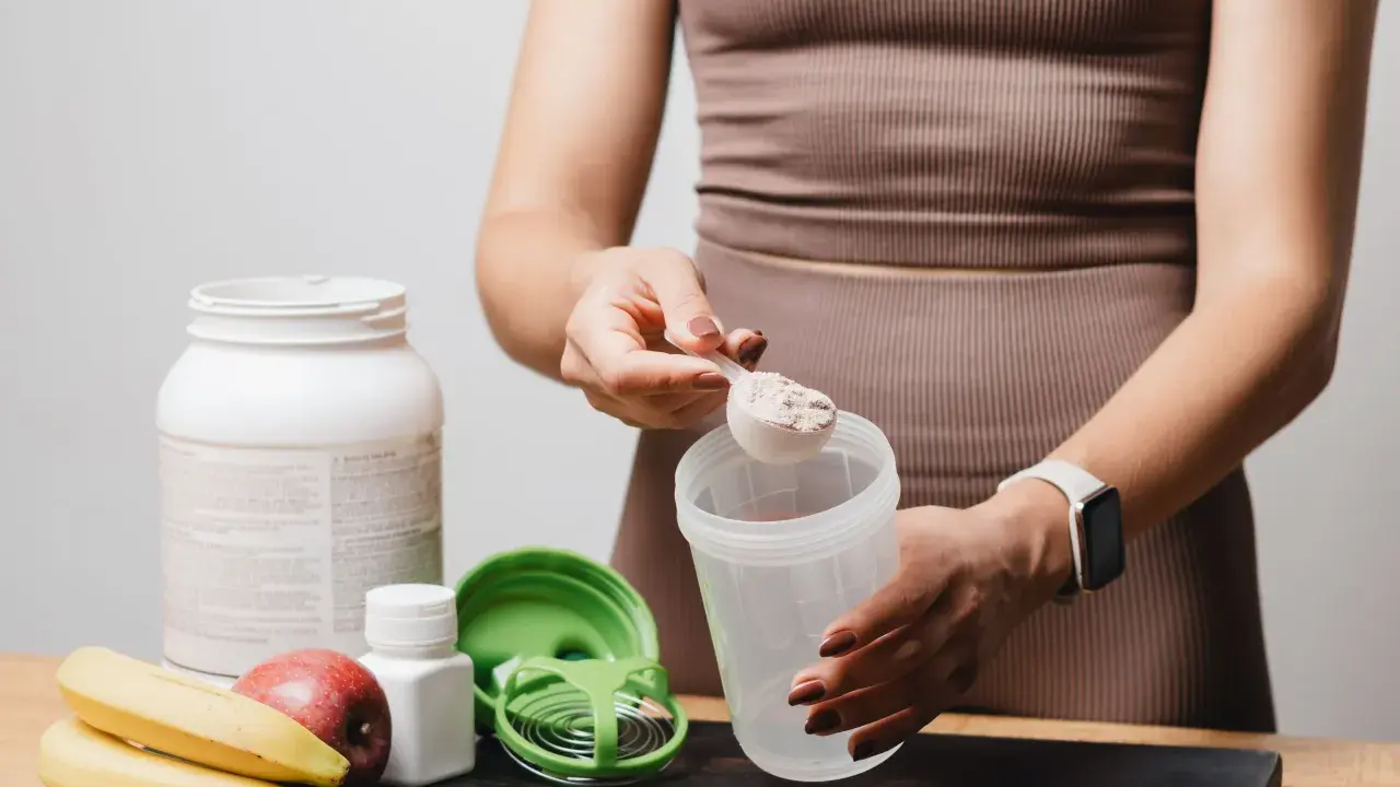 Persona prepara su batido de proteínas. Muestra cómo tomar proteína con una cuchara y un shaker, junto a frutas y suplementos.