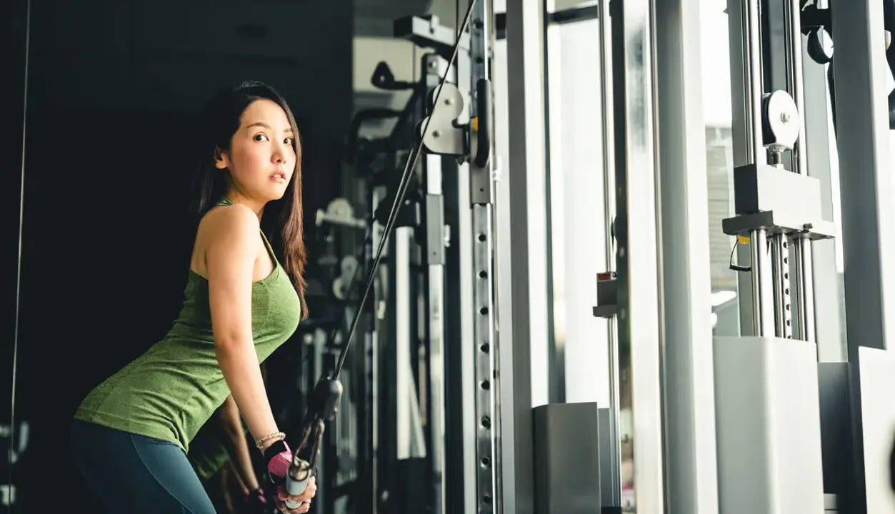 Mujer asiática realiza extensión de triceps en maquina de poleas, concentrada en su entrenamiento.