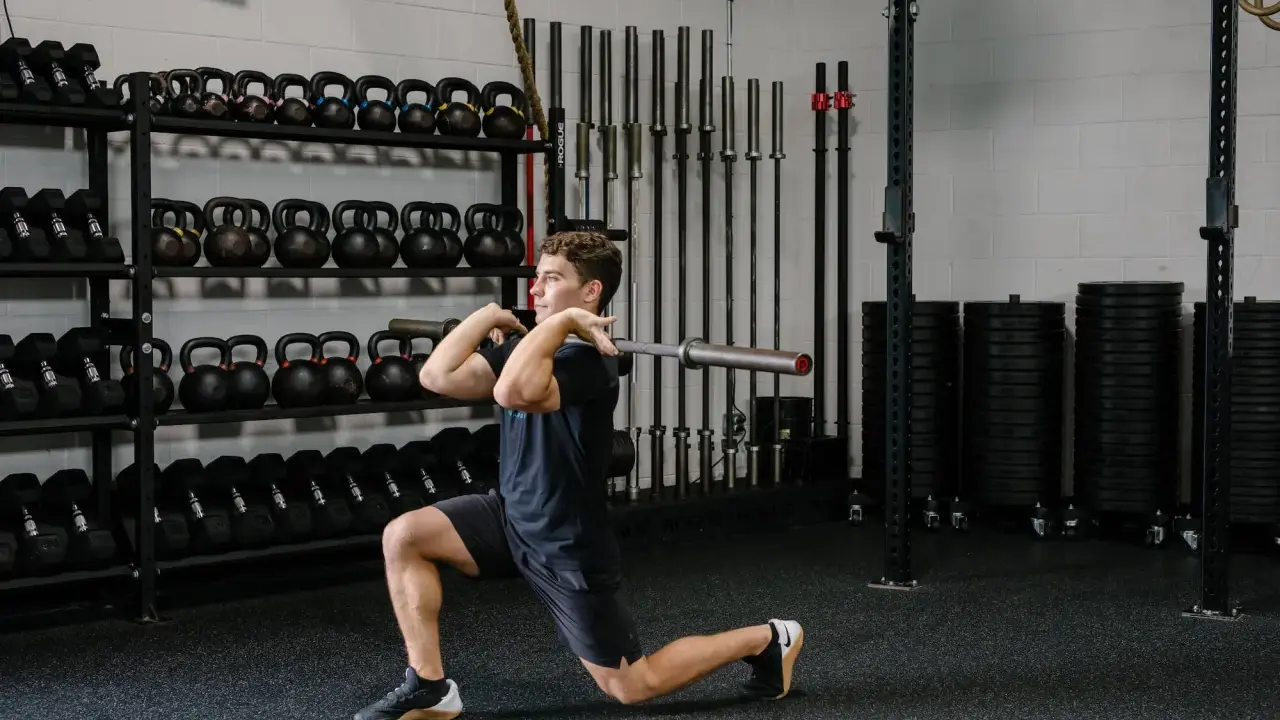 Hombre realizando un front rack lunge con barra en un gimnasio, rodeado de pesas y barras.
