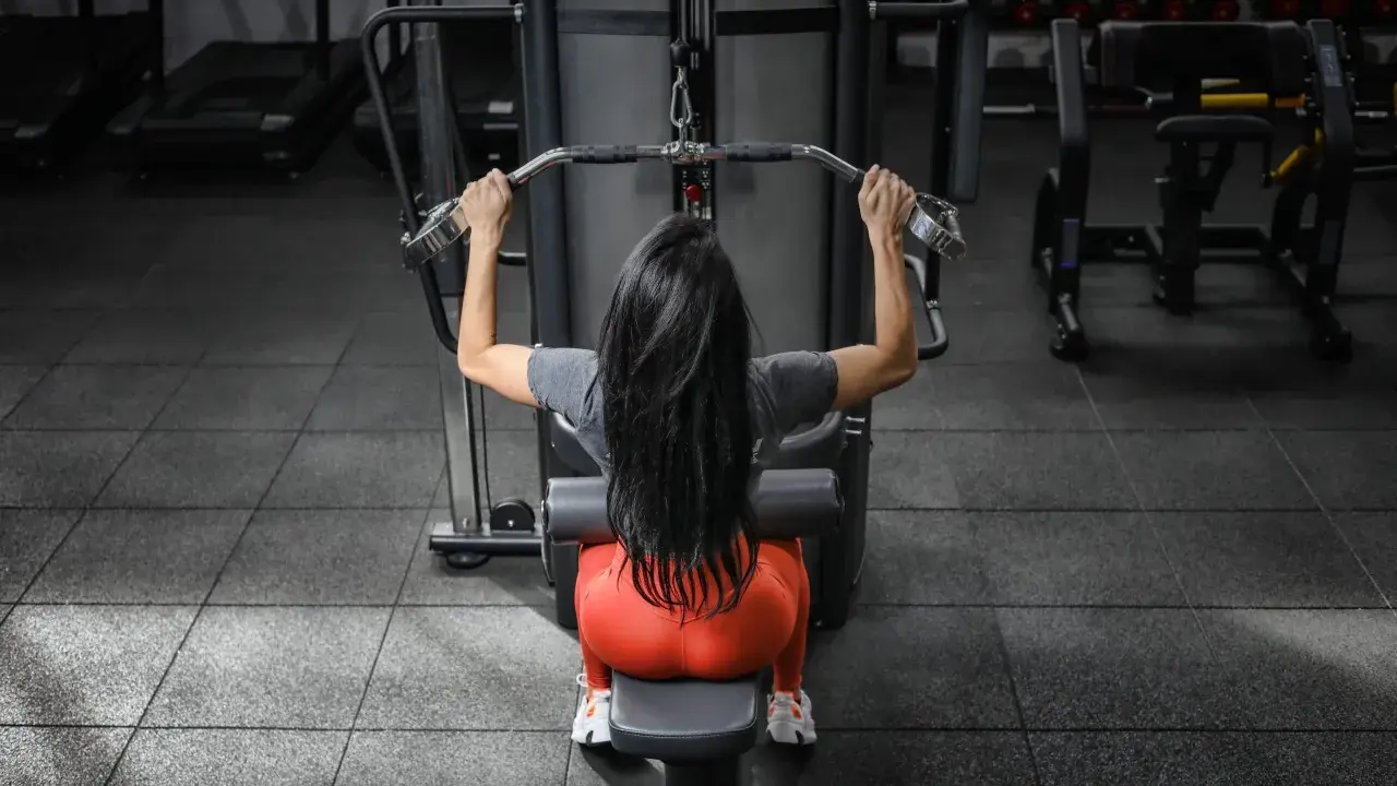 Mujer con cabello largo y negro realiza ejercicios de antebrazo en polea, sentada en una máquina de gimnasio.