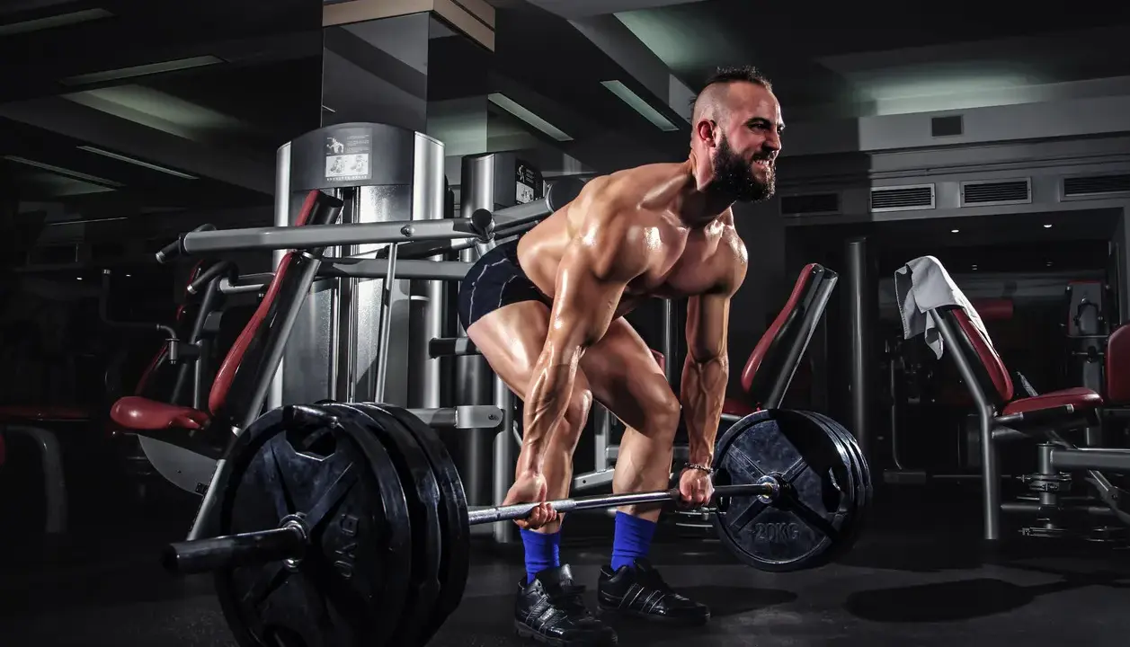 Hombre musculoso levanta una barra con pesas en un gimnasio. Su cuerpo está sudoroso y tenso, listo para el **pes mort**.
