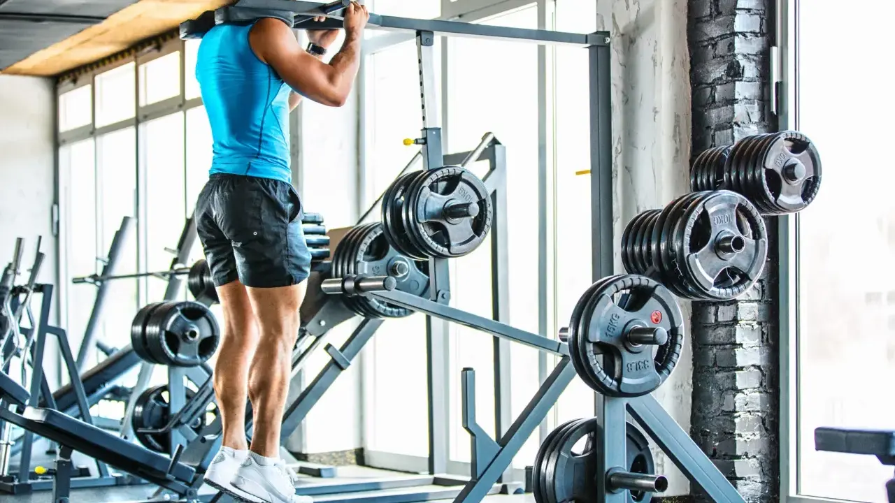 Hombre entrenando en gimnasio, con barra en hombros. Se ven discos de pesas y máquinas.
