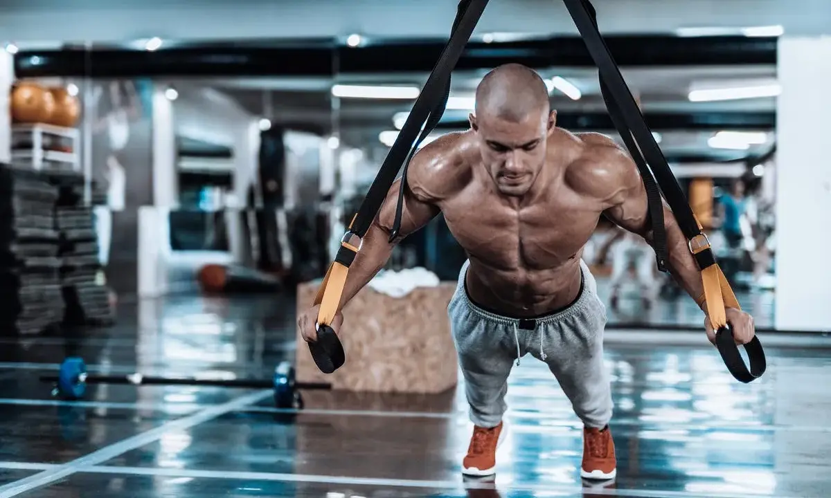 Hombre musculoso realizando trx ejercicios en un gimnasio, con el cuerpo tenso y sudoroso.