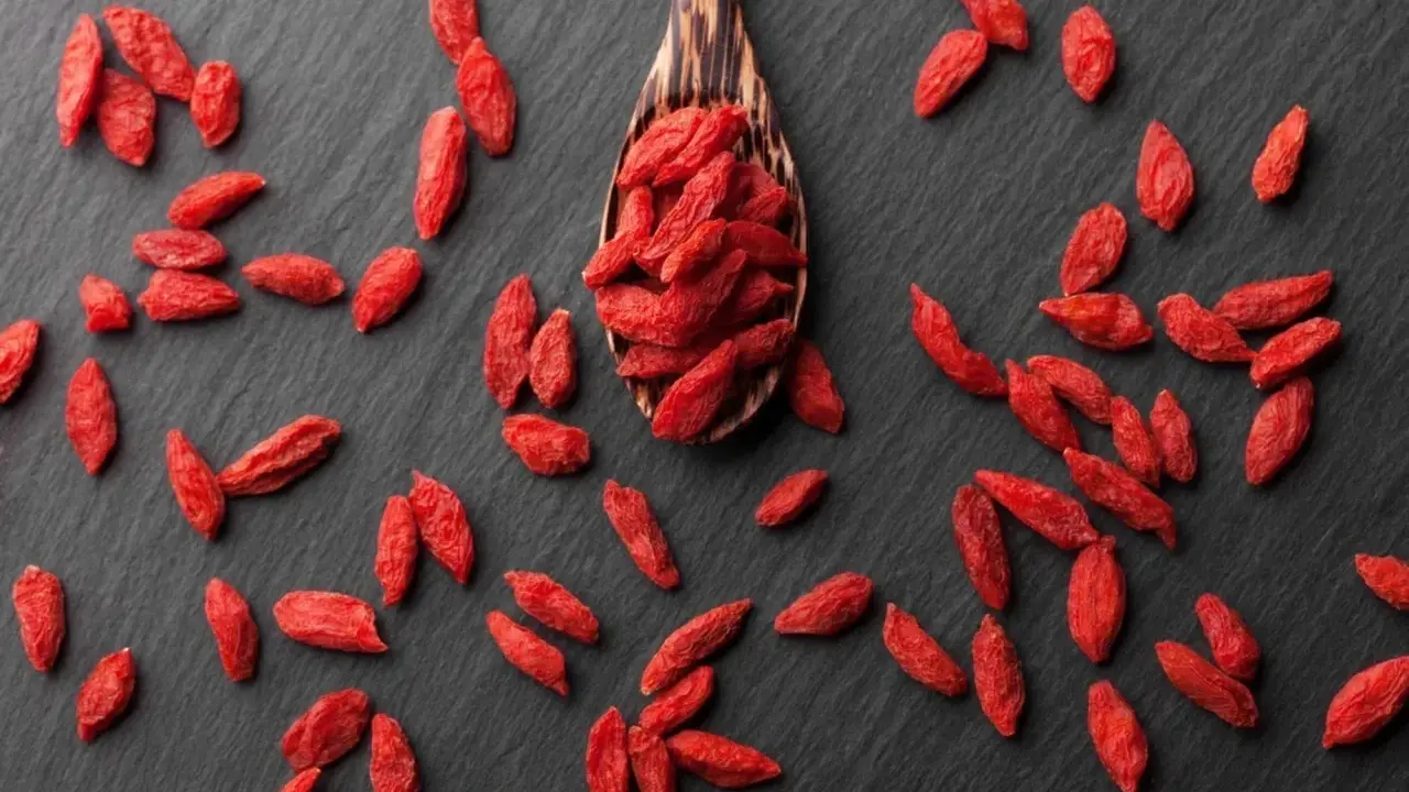 Cucharón de madera lleno de bayas de goji rojas y secas, esparcidas sobre una superficie oscura.
