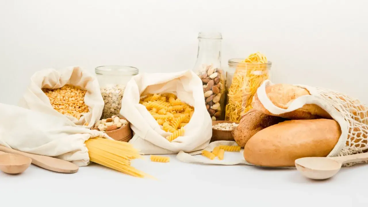 Variedad de alimentos para una carga de hidratos: pasta, pan, avena, frutos secos y legumbres en bolsas de tela y frascos.