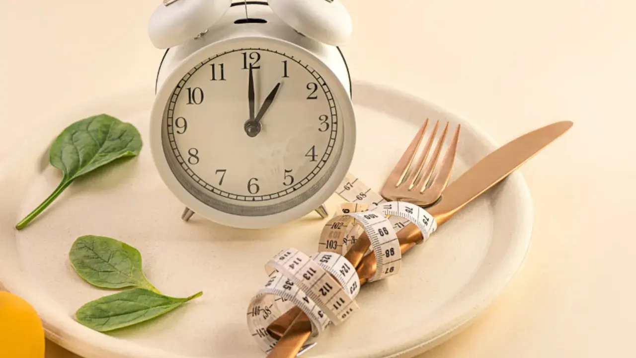 Reloj, plato, cubiertos con cinta métrica y hojas verdes. Símbolos de los beneficios del ayuno intermitente y un estilo de vida saludable.