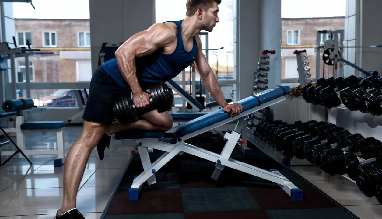 Hombre musculoso realiza remo sentado con mancuerna en gimnasio.