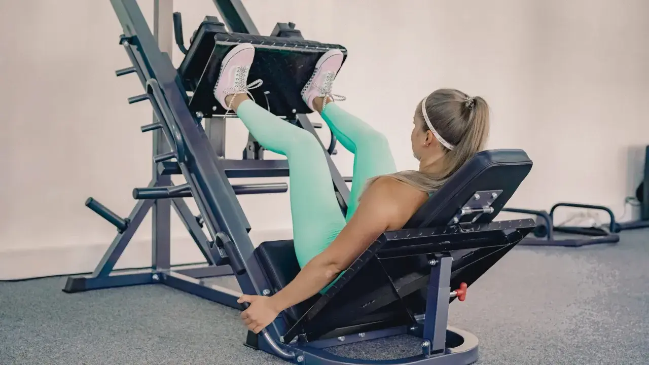 Mujer rubia con leggings turquesa y zapatillas rosas haciendo prensa ejercicio en una máquina de gimnasio.