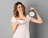 Mujer sonriente con un tazón de cereal y un despertador, lista para el ayuno intermitente para adelgazar 10 kilos.