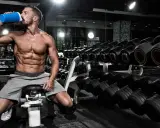 Hombre musculoso bebe de un batido azul en el gimnasio, rodeado de pesas. Es el momento perfecto para la ventana anabolica.