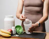 Persona prepara su batido de proteínas. Muestra cómo tomar proteína con una cuchara y un shaker, junto a frutas y suplementos.