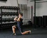 Hombre realizando un front rack lunge con barra en un gimnasio, rodeado de pesas y barras.