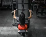 Mujer con cabello largo y negro realiza ejercicios de antebrazo en polea, sentada en una máquina de gimnasio.