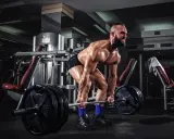 Hombre musculoso levanta una barra con pesas en un gimnasio. Su cuerpo está sudoroso y tenso, listo para el **pes mort**.