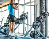 Hombre entrenando en gimnasio, con barra en hombros. Se ven discos de pesas y máquinas.
