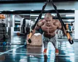 Hombre musculoso realizando trx ejercicios en un gimnasio, con el cuerpo tenso y sudoroso.