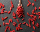 Cucharón de madera lleno de bayas de goji rojas y secas, esparcidas sobre una superficie oscura.