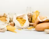 Variedad de alimentos para una carga de hidratos: pasta, pan, avena, frutos secos y legumbres en bolsas de tela y frascos.