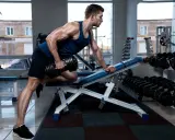Hombre musculoso realiza remo sentado con mancuerna en gimnasio.