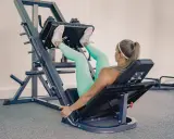 Mujer rubia con leggings turquesa y zapatillas rosas haciendo prensa ejercicio en una máquina de gimnasio.