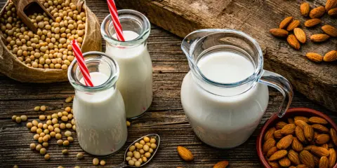 Dos botellas y una jarra de bebida vegetal, junto a un saco de soja y un cuenco de almendras.