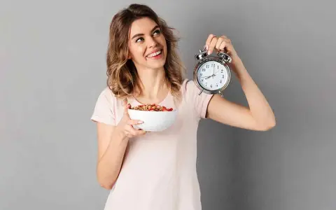 Mujer sonriente con un tazón de cereal y un despertador, lista para el ayuno intermitente para adelgazar 10 kilos.
