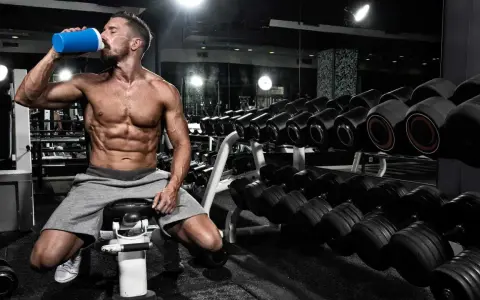 Hombre musculoso bebe de un batido azul en el gimnasio, rodeado de pesas. Es el momento perfecto para la ventana anabolica.