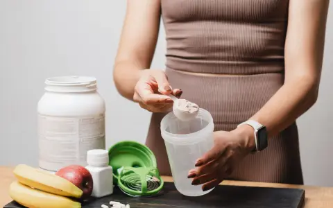 Persona prepara su batido de proteínas. Muestra cómo tomar proteína con una cuchara y un shaker, junto a frutas y suplementos.