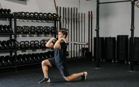 Hombre realizando un front rack lunge con barra en un gimnasio, rodeado de pesas y barras.
