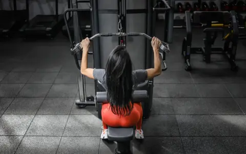 Mujer con cabello largo y negro realiza ejercicios de antebrazo en polea, sentada en una máquina de gimnasio.