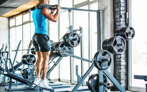Hombre entrenando en gimnasio, con barra en hombros. Se ven discos de pesas y máquinas.
