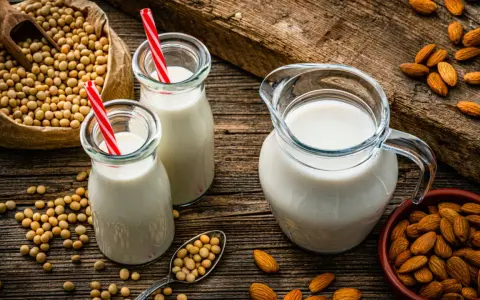 Dos botellas y una jarra de bebida vegetal, junto a un saco de soja y un cuenco de almendras.
