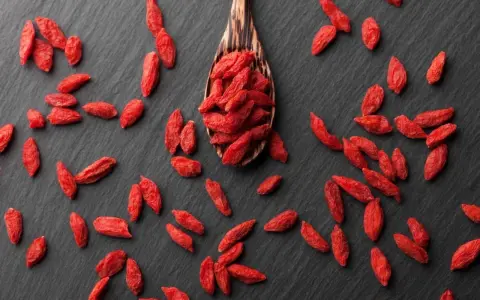 Cucharón de madera lleno de bayas de goji rojas y secas, esparcidas sobre una superficie oscura.