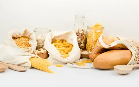 Variedad de alimentos para una carga de hidratos: pasta, pan, avena, frutos secos y legumbres en bolsas de tela y frascos.