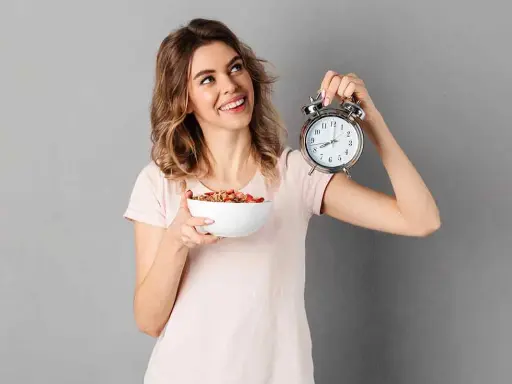 Mujer sonriente con un tazón de cereal y un despertador, lista para el ayuno intermitente para adelgazar 10 kilos.