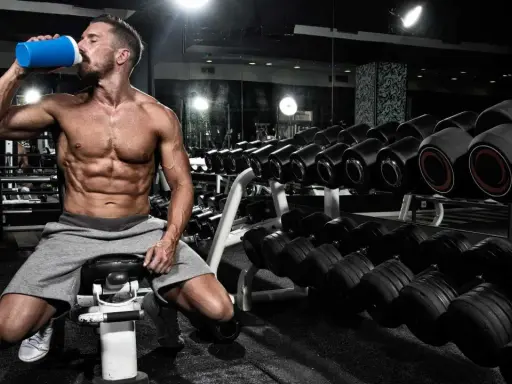 Hombre musculoso bebe de un batido azul en el gimnasio, rodeado de pesas. Es el momento perfecto para la ventana anabolica.