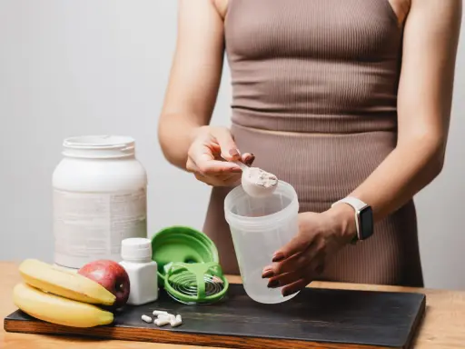 Persona prepara su batido de proteínas. Muestra cómo tomar proteína con una cuchara y un shaker, junto a frutas y suplementos.
