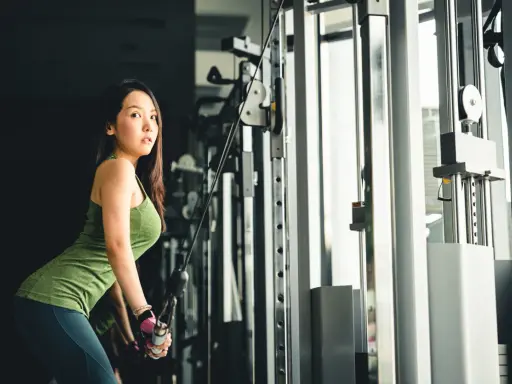 Mujer asiática realiza extensión de triceps en maquina de poleas, concentrada en su entrenamiento.