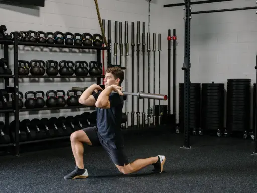 Hombre realizando un front rack lunge con barra en un gimnasio, rodeado de pesas y barras.