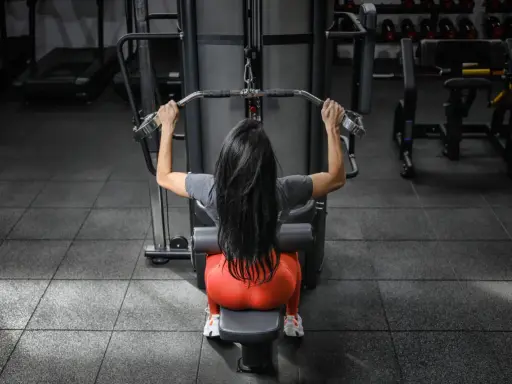 Mujer con cabello largo y negro realiza ejercicios de antebrazo en polea, sentada en una máquina de gimnasio.