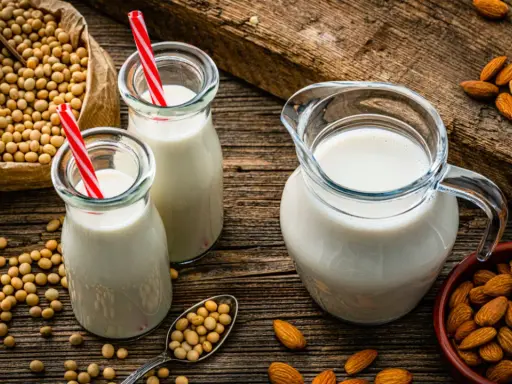 Dos botellas y una jarra de bebida vegetal, junto a un saco de soja y un cuenco de almendras.
