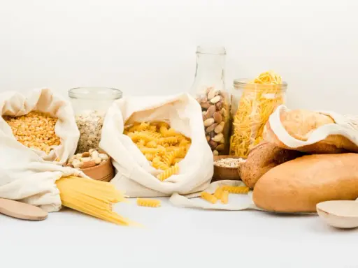 Variedad de alimentos para una carga de hidratos: pasta, pan, avena, frutos secos y legumbres en bolsas de tela y frascos.