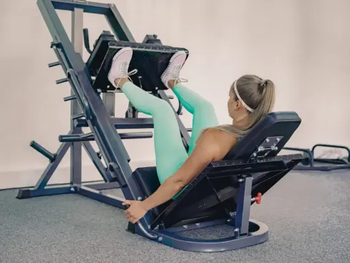 Mujer rubia con leggings turquesa y zapatillas rosas haciendo prensa ejercicio en una máquina de gimnasio.