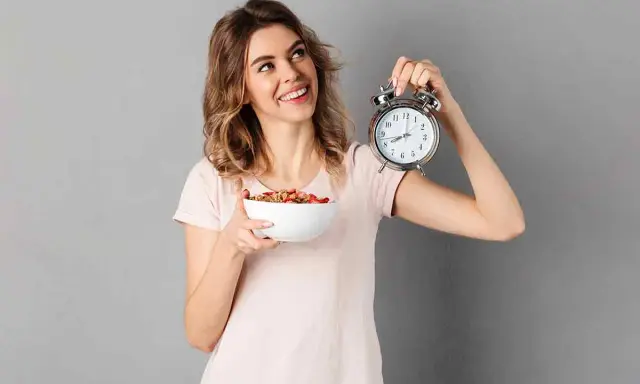 Mujer sonriente con un tazón de cereal y un despertador, lista para el ayuno intermitente para adelgazar 10 kilos.