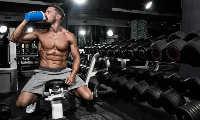 Hombre musculoso bebe de un batido azul en el gimnasio, rodeado de pesas. Es el momento perfecto para la ventana anabolica.