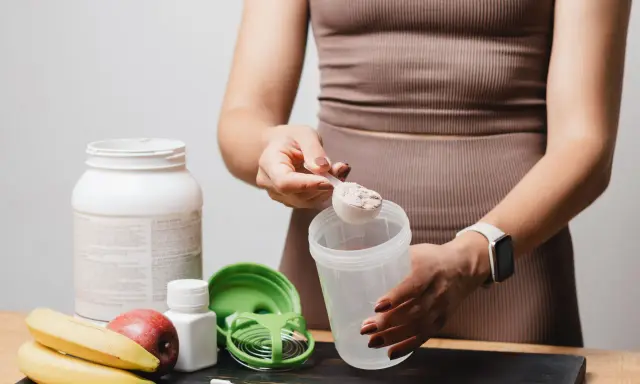 Persona prepara su batido de proteínas. Muestra cómo tomar proteína con una cuchara y un shaker, junto a frutas y suplementos.