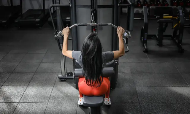 Mujer con cabello largo y negro realiza ejercicios de antebrazo en polea, sentada en una máquina de gimnasio.