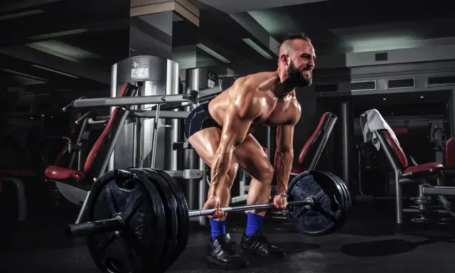 Hombre musculoso levanta una barra con pesas en un gimnasio. Su cuerpo está sudoroso y tenso, listo para el **pes mort**.